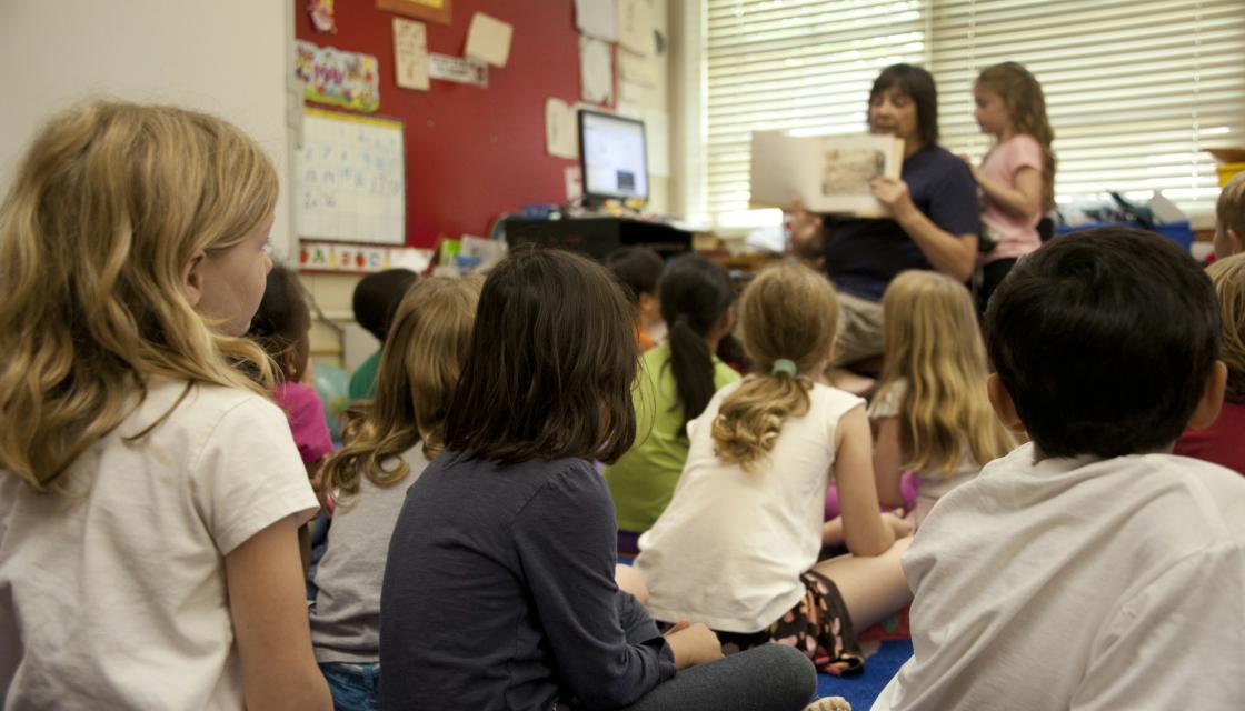 A group of children in a classroom with a teacher holding up a book