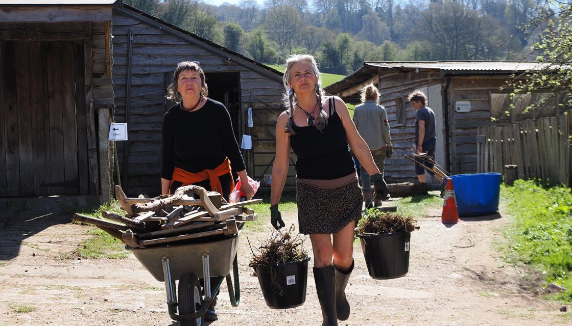 Two women in a farming setting, one with a wheelbarrow, the other carrying buckets