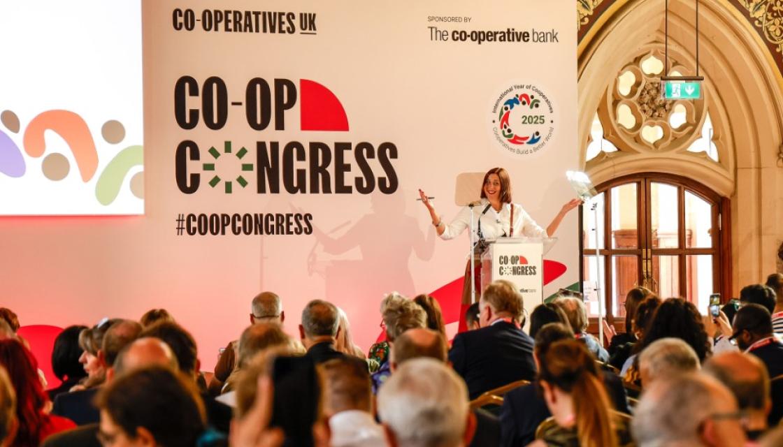 A photograph of Rose Marley, on stage at Co-op Congress 2025, addressing a packed out Rochdale Town Hall.