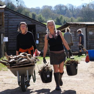 Two women in a farming setting, one with a wheelbarrow, the other carrying buckets
